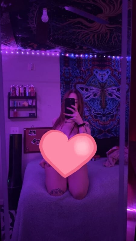 Young woman with long hair sitting on a bed in a dimly lit room with purple lighting, tapestries, and neon decorations on the wall.