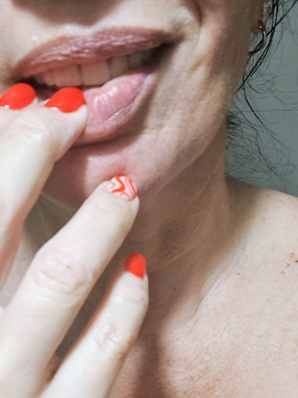 Close-up of a woman with red nail polish touching her lips and teeth, smiling slightly.