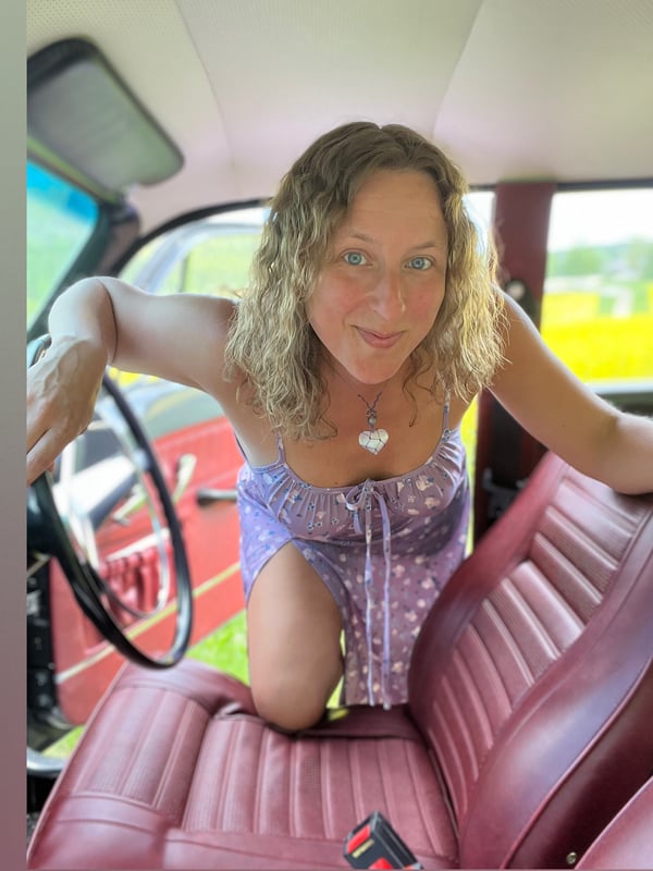 A smiling woman with wavy blonde hair leans forward in the driver's seat of a vintage red car, wearing a purple dress.