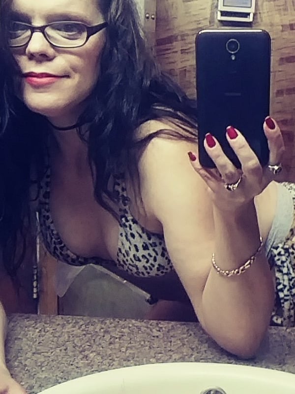 Woman taking a selfie in a bathroom mirror, wearing glasses, a choker, and leopard print bikini while holding her phone.