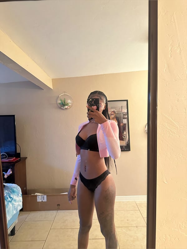 A woman takes a mirror selfie in a bedroom, wearing a black bikini top with a pink sheer cover-up, standing confidently.