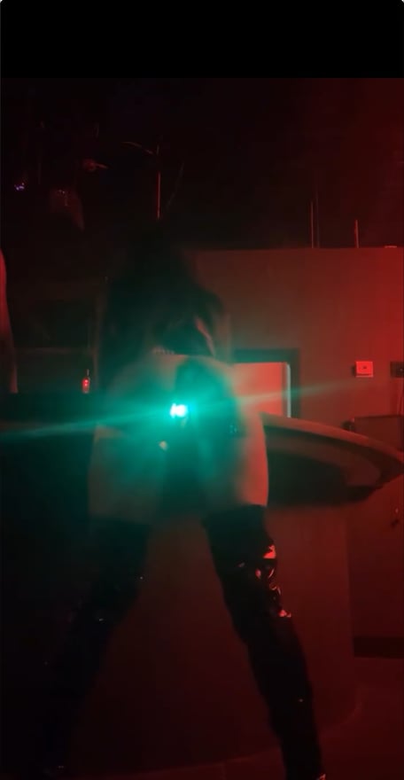 A person standing in shiny boots in a dimly lit room with red lighting and a green light effect.