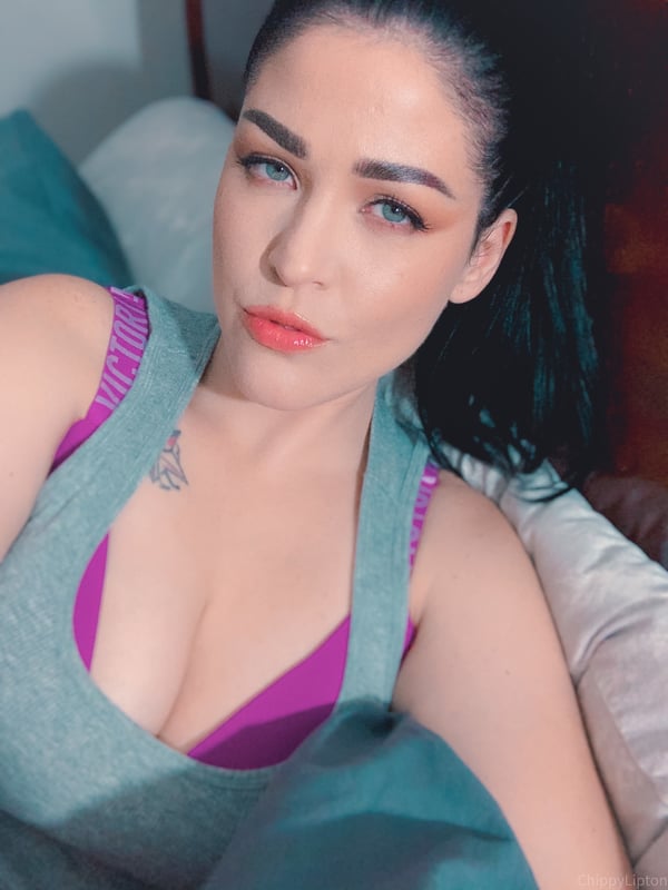 A woman with dark hair in a ponytail sitting on a bed, wearing a gray top with purple straps, smiling at the camera.