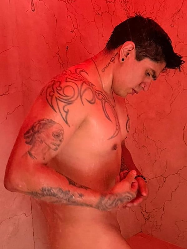Young man with tattoos and piercings posing shirtless in a red-lit room.