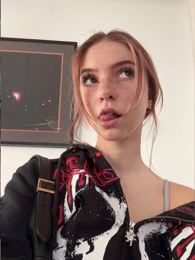 A young woman with brown hair poses indoors wearing a graphic top and jacket.