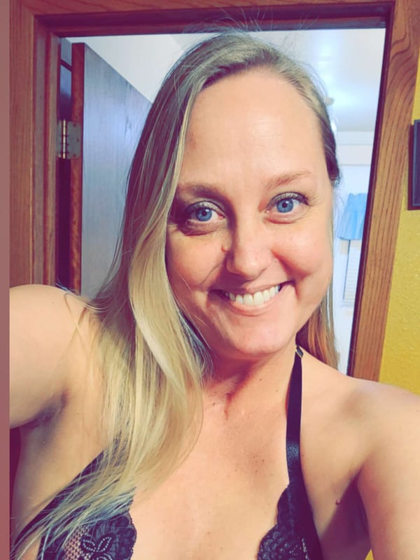 A smiling woman with blonde hair taking a selfie in a doorway, wearing black lace lingerie.