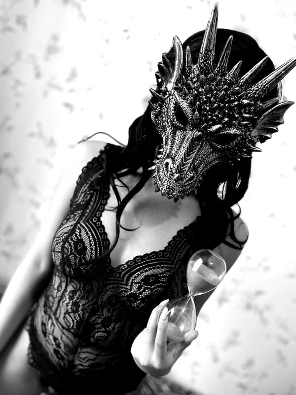 A woman wearing an elaborate dragon mask and lace lingerie, holding an hourglass, posed dramatically in black and white.