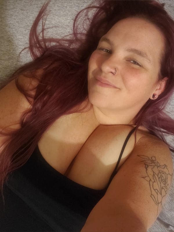 A smiling woman with long reddish-purple hair lying on a bed in a black tank top, showing a tattoo on her arm.