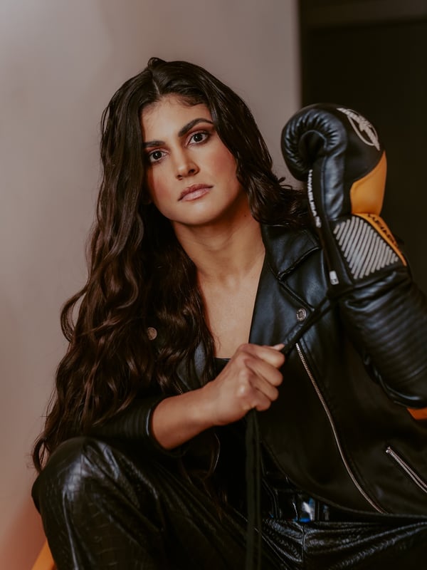 A woman with long dark hair posing with boxing gloves and wearing a leather jacket and pants in a studio setting.