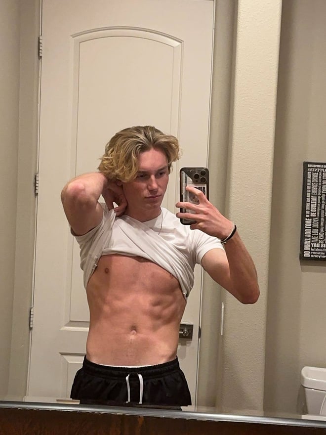 Young man with curly blonde hair taking a mirror selfie in a locker room, wearing a white t-shirt and grey sweatpants.