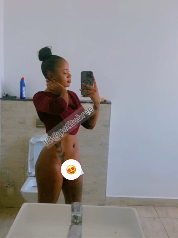Young woman with dark hair in a bun taking a mirror selfie in a bathroom, wearing a maroon off-shoulder top and posing confidently.