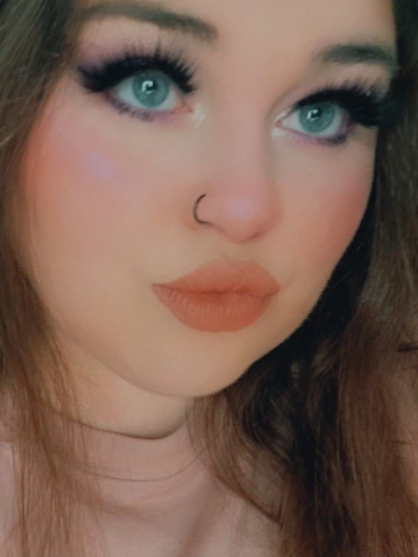 Close-up portrait of a young woman with long brown hair, green eyes, and makeup, wearing a subtle nose piercing.