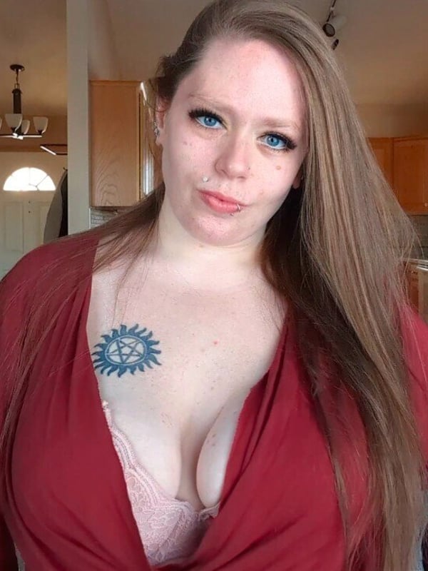 Woman with long hair posing in a kitchen, wearing a red cardigan over a pink top with a visible tattoo on her chest.