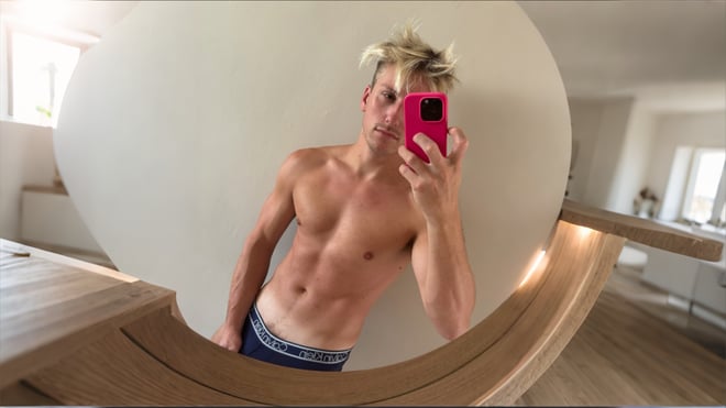 A fit blond man taking a mirror selfie in a bright modern room.