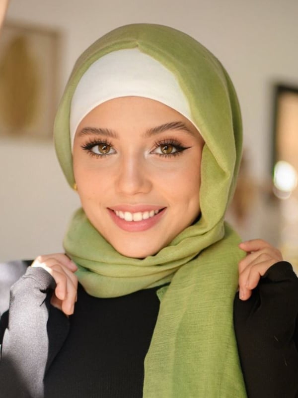 Young woman smiling with green hijab and black top, hands on shoulders, against a simple indoor background with curtains.