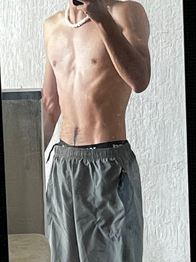 A slim person with a pearl necklace posing shirtless in front of a mirror, wearing gray shorts.