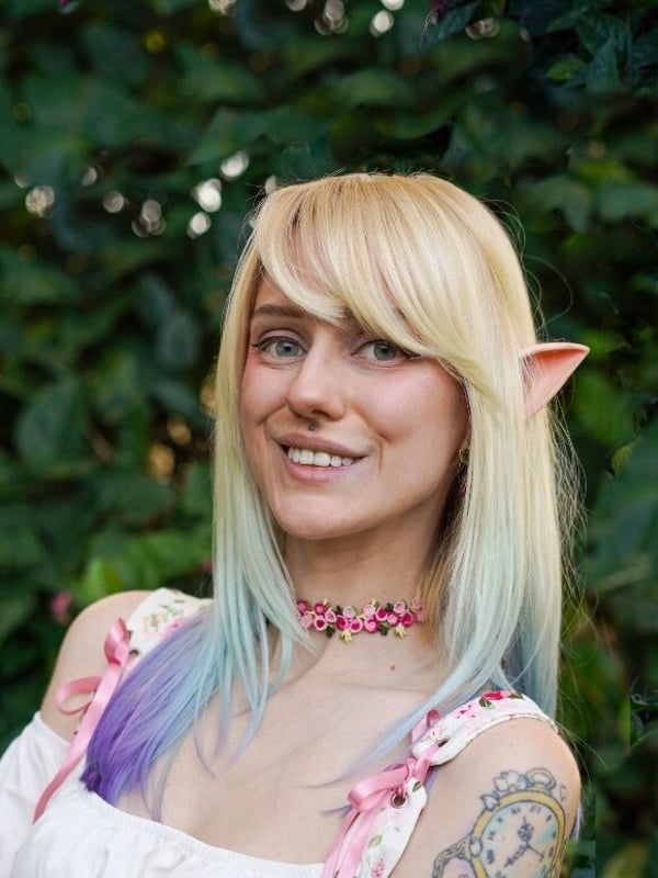 Woman with long blonde hair dyed with blue and purple tips, wearing elf ears and a floral choker, smiling against a green outdoor background.
