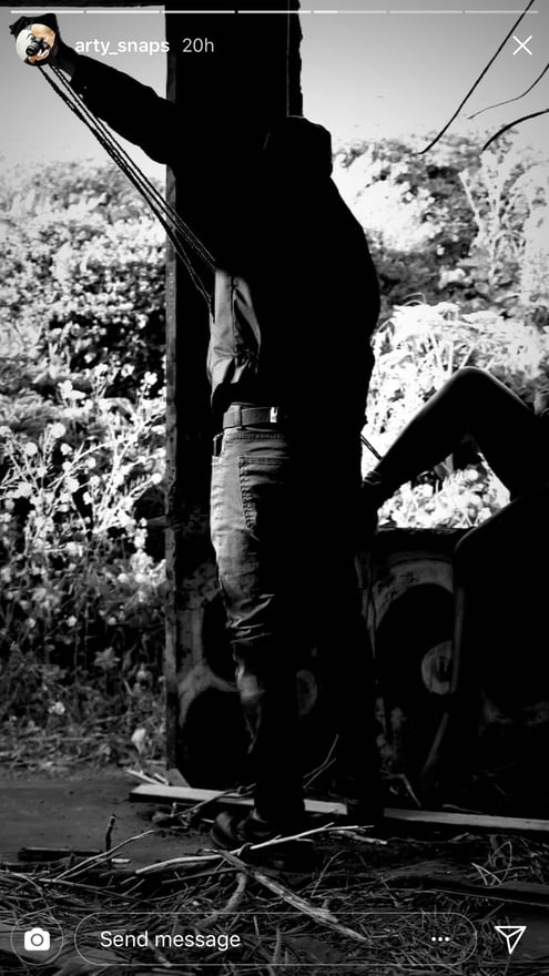 A person in dark clothing posing dynamically near a tree and plants in an outdoor setting with a vintage filter.