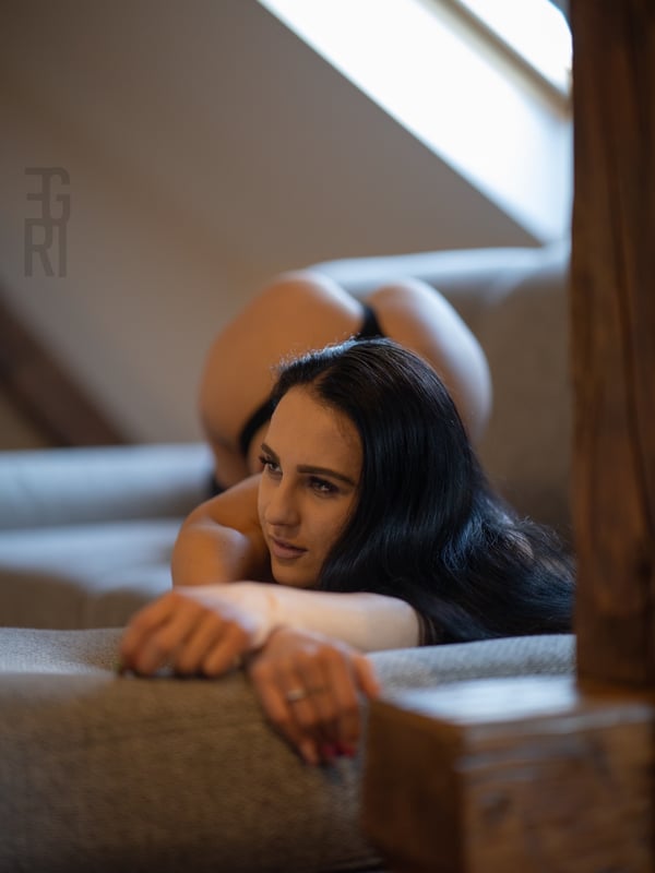 A woman posing on a couch near a window, leaning forward with long dark hair.