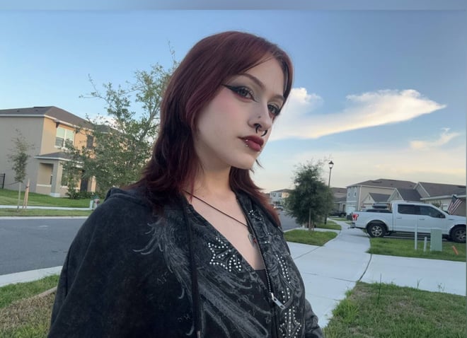 A red-haired woman with facial piercings posing outdoors in a suburban neighborhood.
