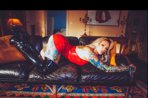 A woman with blonde hair and tattoos poses seductively on a leather couch in a red dress and black boots, in a warmly lit living room with vintage decor.