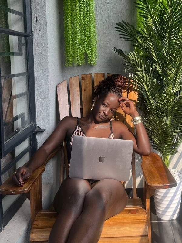 A woman with curly hair sits relaxed on a wooden chair on a balcony surrounded by plants, holding a laptop.