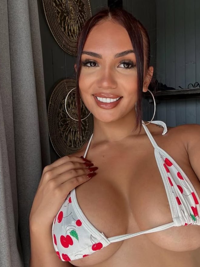A smiling woman with long hair and hoop earrings poses in a cherry-patterned bikini top indoors near curtains and decor.