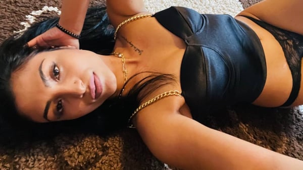 A woman with long dark hair lying on a rug, wearing black lingerie and jewelry, posing seductively.
