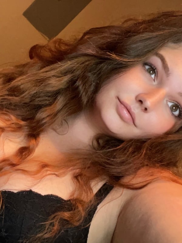 Close-up selfie of a young woman with wavy red hair, wearing black lingerie, smiling softly.