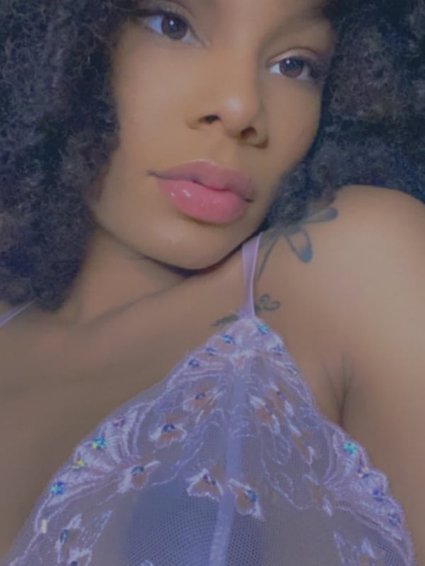 Young woman with curly hair posing in a sheer top indoors, smiling at the camera with tattoos visible on her arms.