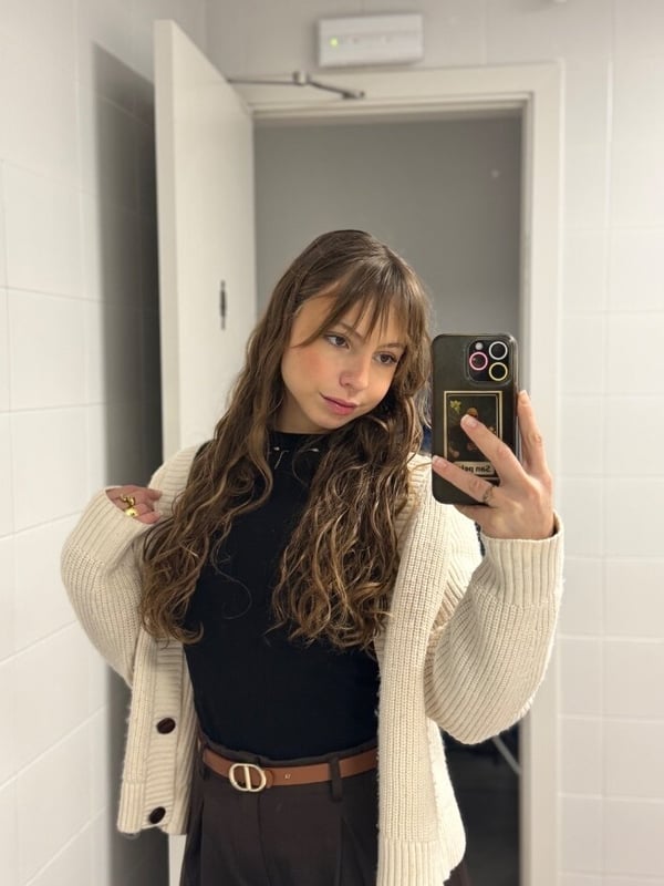 Young woman with long wavy brown hair taking a mirror selfie in a bathroom, wearing a white cardigan over a black top and brown skirt.