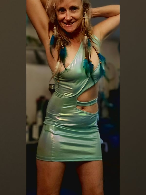 A woman with long blonde hair raising her arms in a shiny green dress, posing indoors with a playful expression.