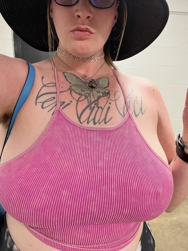 A woman taking a selfie in a pink tank top and wide-brim hat, showing tattoos on her chest and arm, with a necklace and wristwatch.