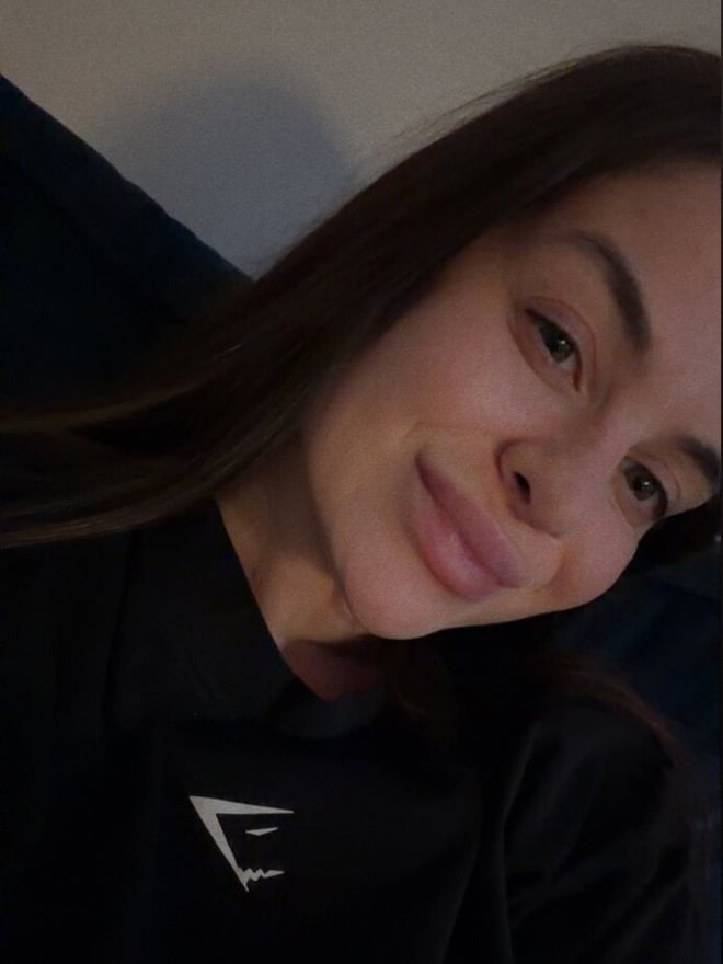A young woman with long brown hair smiling softly while reclining on a cushioned surface, wearing a black athletic top.