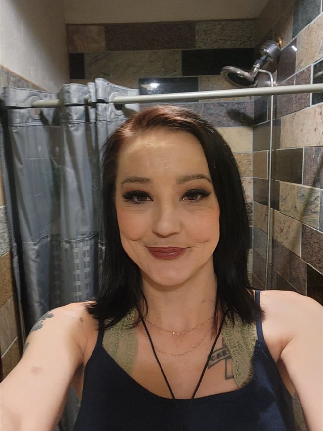 A woman with shoulder-length dark hair and tattoos on her arm takes a selfie in a bathroom with tiled walls and a shower curtain in the background.