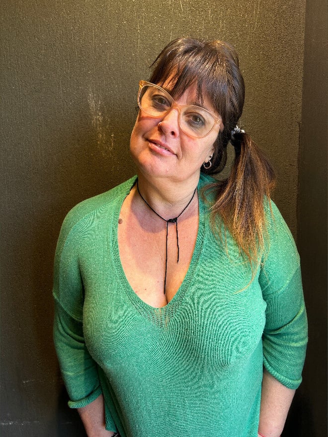 A woman with glasses and ponytail smiling at the camera, wearing a green sweater with a choker necklace.