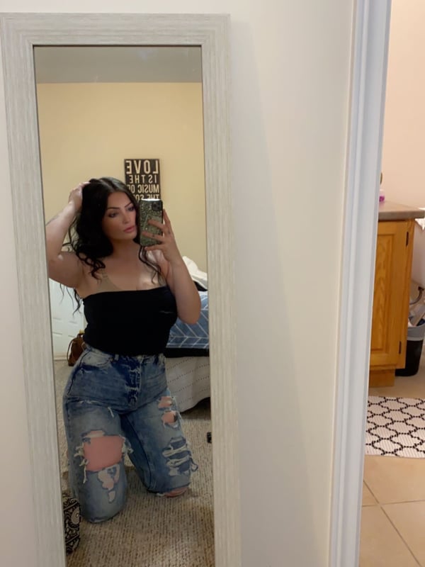 A woman taking a mirror selfie in a bedroom, wearing a black tube top and ripped jeans while kneeling.