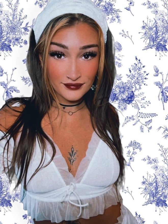 Young woman with blonde-highlighted hair wearing a white headband and lace top, smiling confidently against a floral blue background with choker necklace.