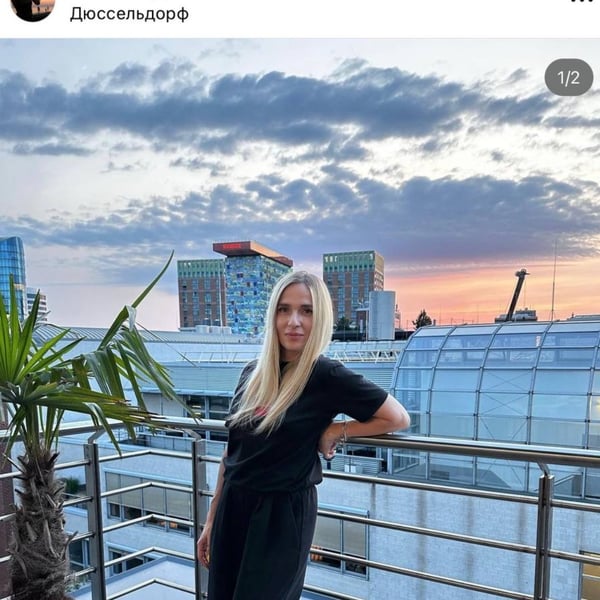A woman with long blonde hair stands on a balcony, posing casually in black clothing against a sunset skyline with modern buildings.