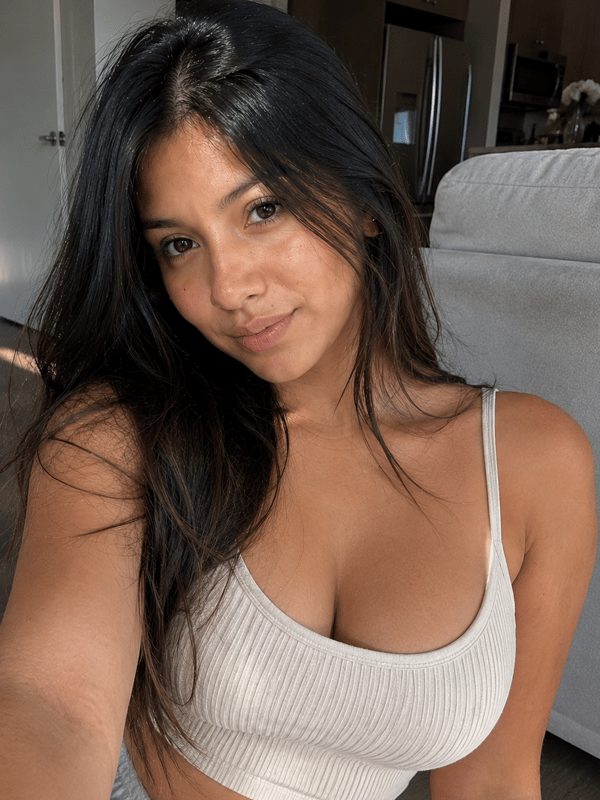 A young woman with long dark hair posing topless while covering her chest with both hands in a bright indoor setting.