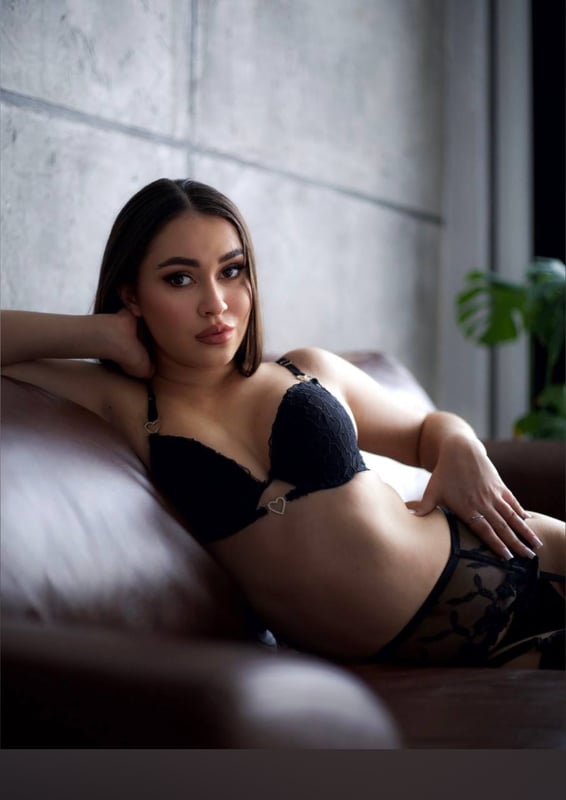 A woman with long dark hair poses seductively on a brown couch against a concrete wall with a plant in the background.