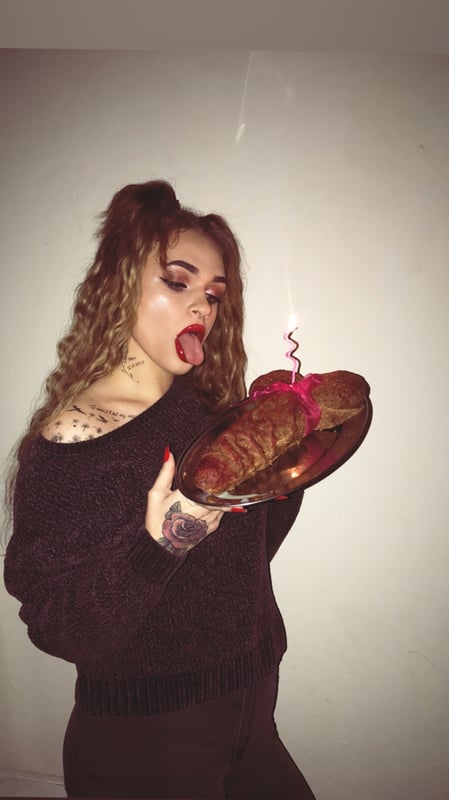 Young woman with curly hair in pigtails, wearing a sweater, playfully holding a plate of pastries with a candle next to a plain wall.