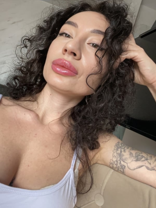 A woman with curly hair and plump lips posing confidently in a white tank top, showing a tattoo on her arm.