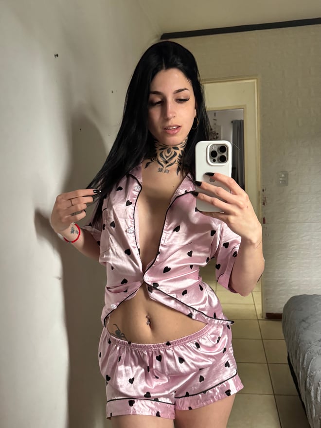 A woman with long dark hair and visible tattoos poses in a red outfit on a carpeted floor.