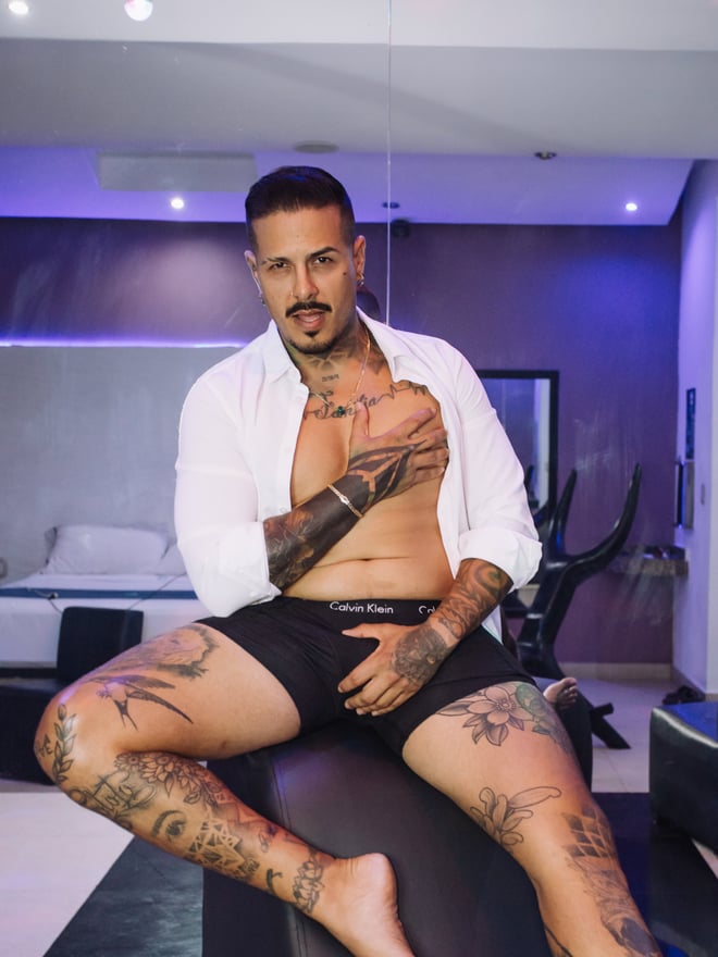 Man sitting on a stool in a modern room with exercise bike, wearing an open white shirt and shorts, displaying tattoos on his chest, arms, and legs.
