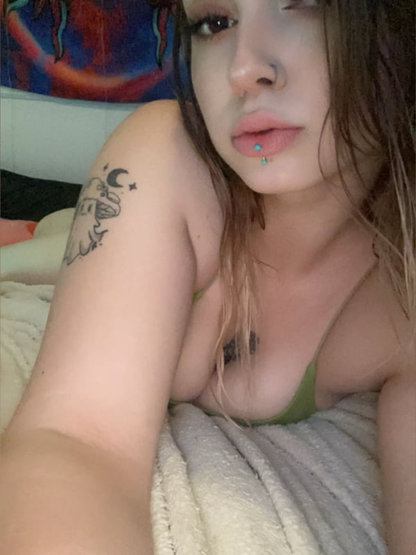 A close-up of a person with distinctive makeup and piercings lying on a bed against a colorful wall hanging, wearing a green top.