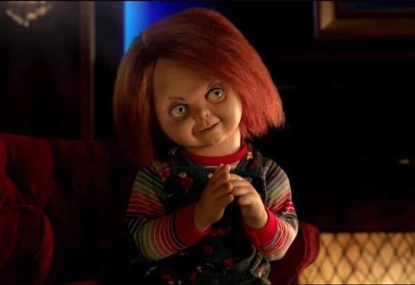Chucky doll from the horror movie sitting on a red chair in a dimly lit room with a blue glowing background.