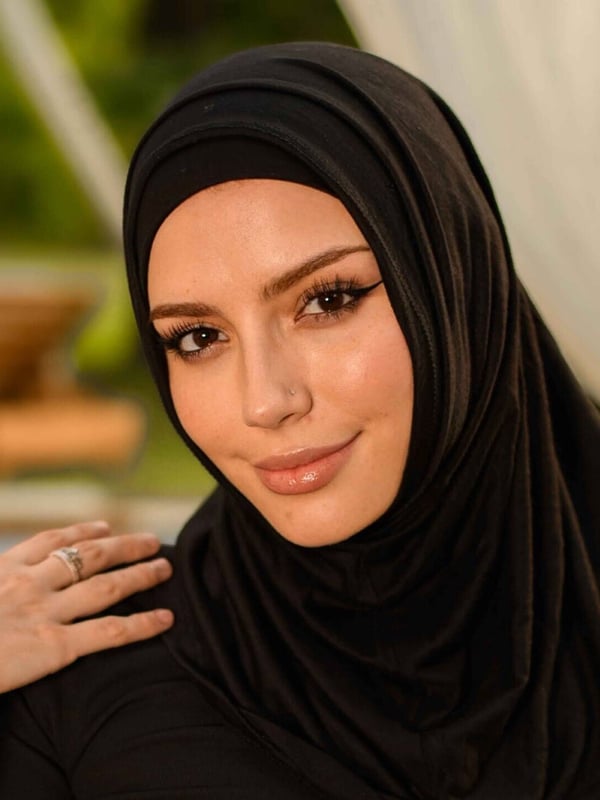 Close-up portrait of a smiling woman wearing a black hijab against a poolside background.
