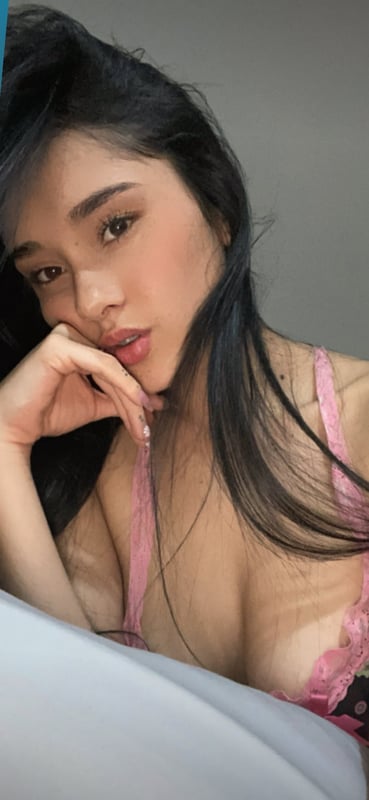 Young woman with long dark hair posing thoughtfully in pink lingerie against a plain background.
