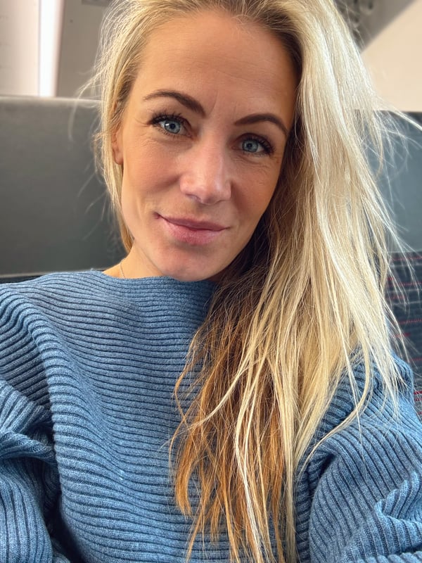 A blonde woman with blue eyes smiling at the camera, wearing a blue knit sweater, seated indoors.